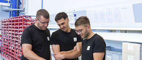 Three men standing in a production hall, jointly looking at a notebook.
