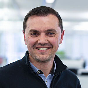 A man in a half-zip sweatshirt stands smiling, looking toward the camera.