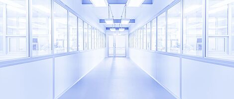 Heller weißer Flur mit einer Glastür am Ende. Links und rechts befinden sich Fenster, hinter denen Personen erkennbar sind. Die Umgebung wirkt steril.