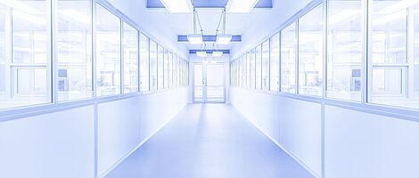 Bright white corridor with a glass door at the end. Windows on both sides reveal visible people behind them. The environment appears sterile.