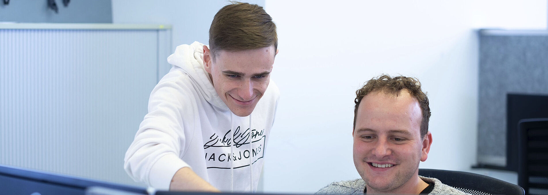 Ein junger Mann sitzt am PC vor zwei Monitoren; neben ihm steht ein weiterer junger Mann und zeigt auf einen der Bildschirme.