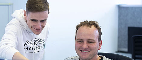 A young man is sitting at a computer with two monitors in front of him; next to him, another young man is standing and pointing at one of the screens.