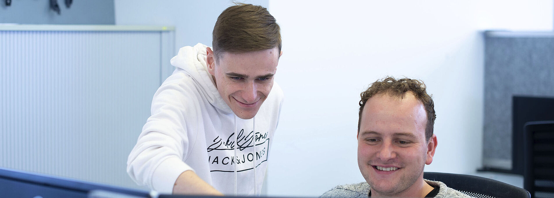 A young man is sitting at a computer with two monitors in front of him; next to him, another young man is standing and pointing at one of the screens.