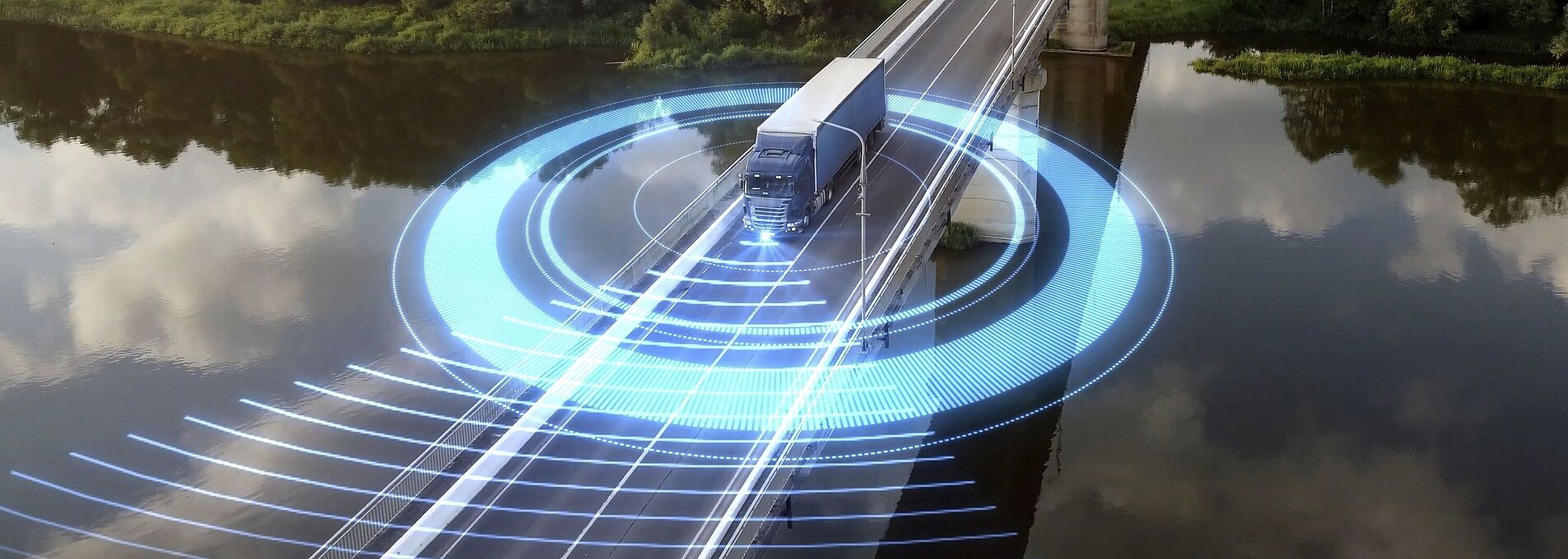 LKW fährt über eine Brücke, die einen Fluss überspannt. Im Hintergrund erstreckt sich ein dichter Wald. Um den LKW kreisen blaue, futuristisch leuchtende Lichter.