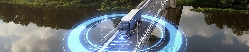 LKW fährt über eine Brücke, die einen Fluss überspannt. Im Hintergrund erstreckt sich ein dichter Wald. Um den LKW kreisen blaue, futuristisch leuchtende Lichter.