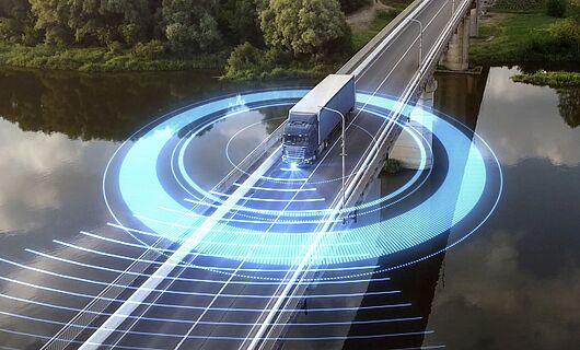 LKW fährt über eine Brücke, die einen Fluss überspannt. Im Hintergrund erstreckt sich ein dichter Wald. Um den LKW kreisen blaue, futuristisch leuchtende Lichter.