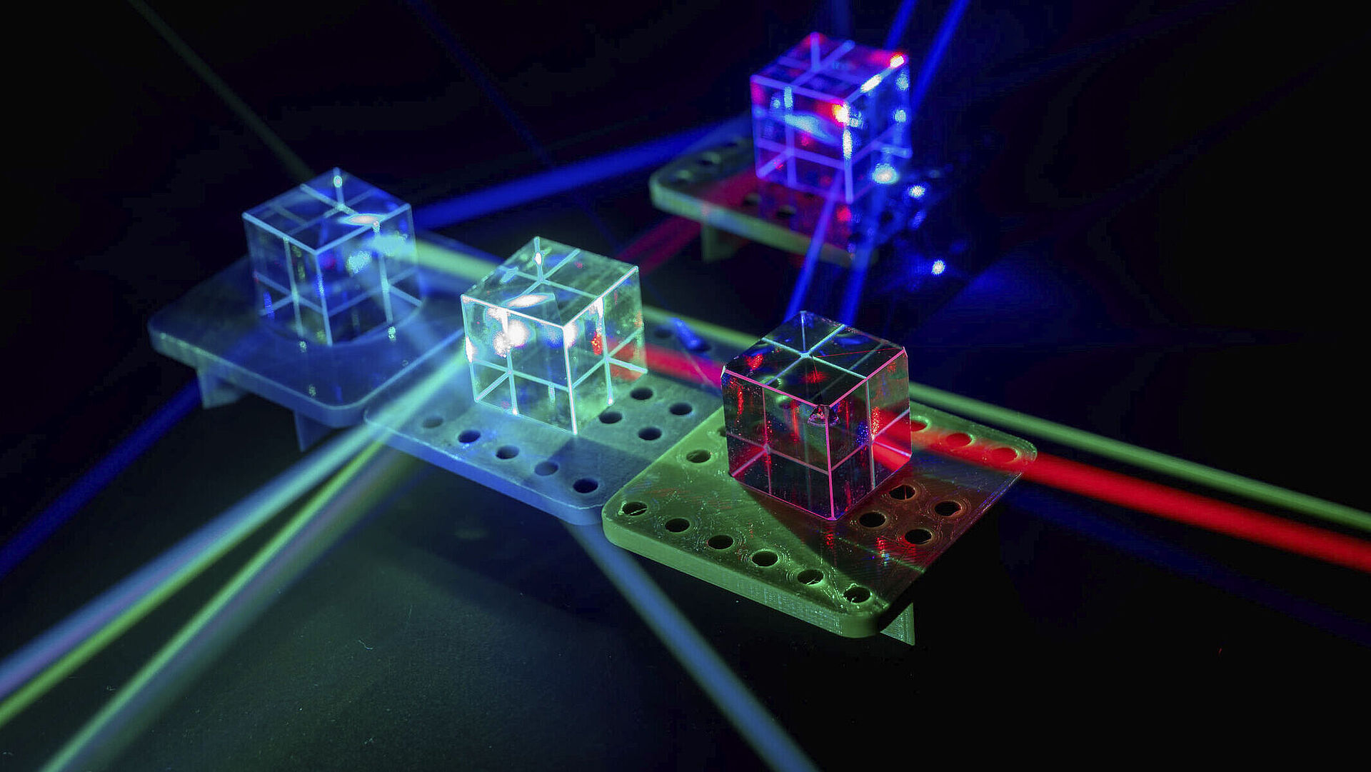 Vier transparente Würfel auf Plateaus, durch die mehrere bunte Laserstrahlen reflektiert werden.
