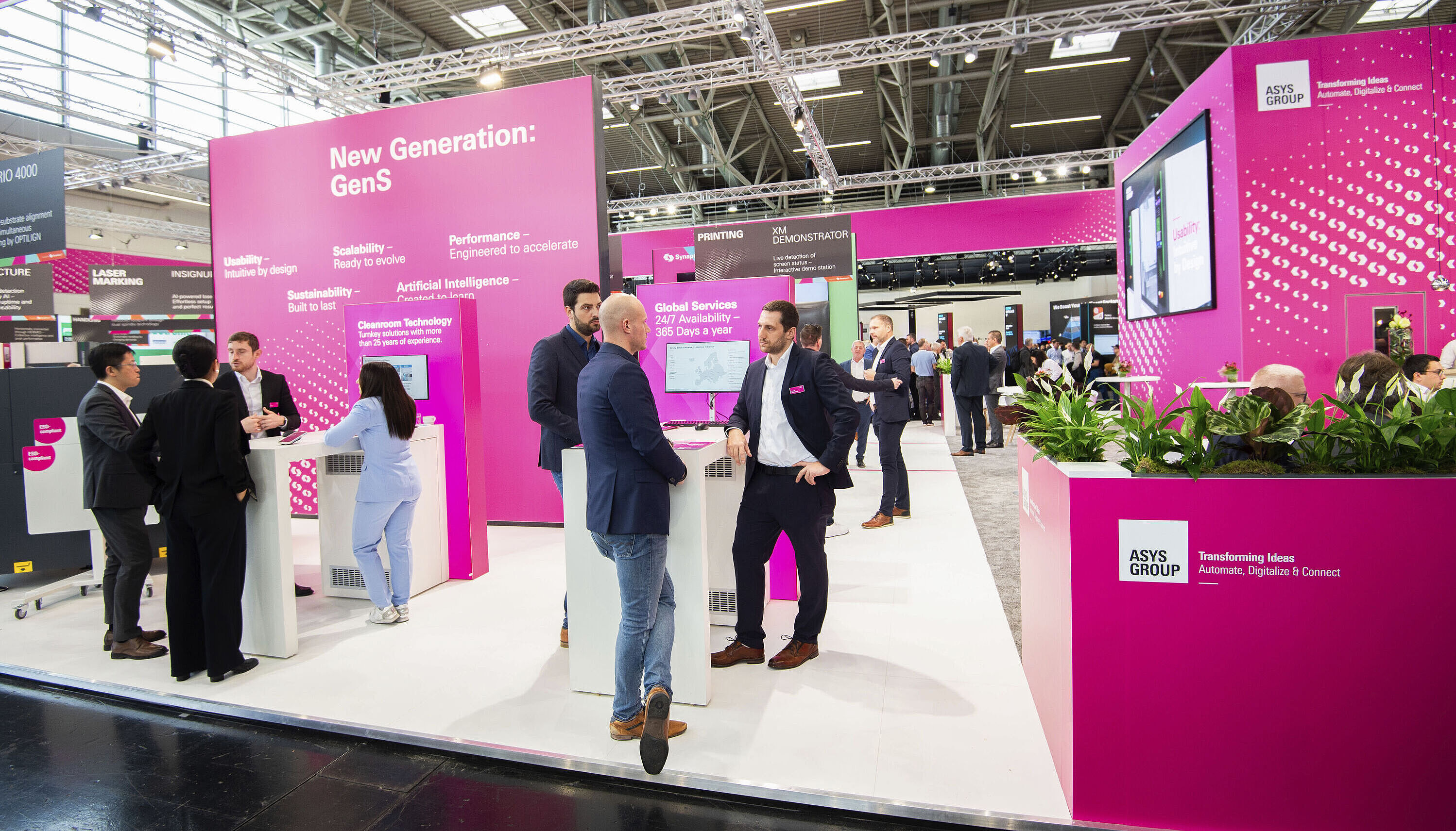 Mehrere Männer und eine Frau in Anzügen auf einem magenta farbenen Messestand. Stehend an Stehtischen und unterhalten sich.