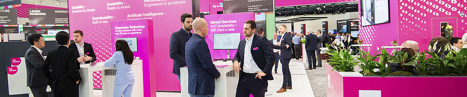 Mehrere Männer und eine Frau in Anzügen auf einem magenta farbenen Messestand. Stehend an Stehtischen und unterhalten sich.