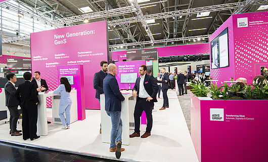 Mehrere Männer und eine Frau in Anzügen auf einem magenta farbenen Messestand. Stehend an Stehtischen und unterhalten sich.