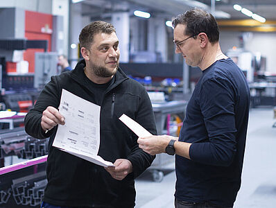 Zwei Männer diskutieren in einer Produktionshalle; einer von ihnen hält einen Plan in der Hand.