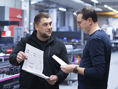 Two men are having a discussion in a production hall; one of them is holding a plan.