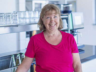 Smiling woman in a magenta T-shirt standing in front of a kitchen counter with many empty glasses.
