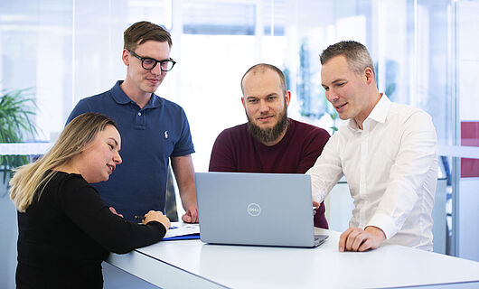 Three men and one woman are standing together at a standing table, looking at a laptop placed on the table.