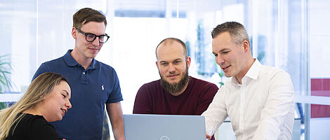 Three men and one woman are standing together at a standing table, looking at a laptop placed on the table.