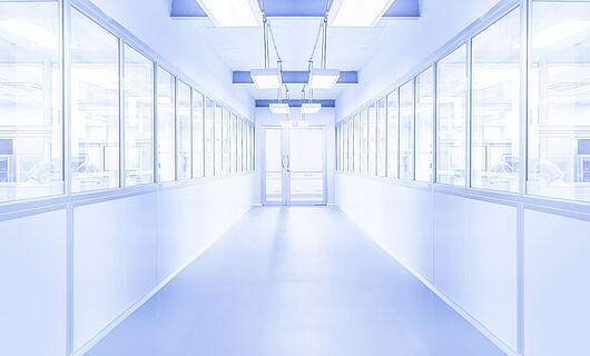 Bright white corridor with a glass door at the end. Windows on both sides reveal visible people behind them. The environment appears sterile.