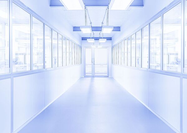 Bright white corridor with a glass door at the end. Windows on both sides reveal visible people behind them. The environment appears sterile.