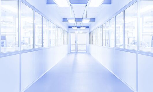 Heller weißer Flur mit einer Glastür am Ende. Links und rechts befinden sich Fenster, hinter denen Personen erkennbar sind. Die Umgebung wirkt steril.