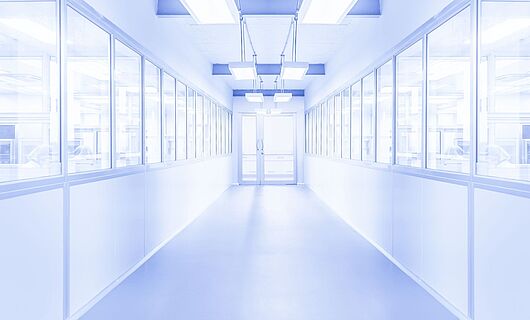 Heller weißer Flur mit einer Glastür am Ende. Links und rechts befinden sich Fenster, hinter denen Personen erkennbar sind. Die Umgebung wirkt steril.