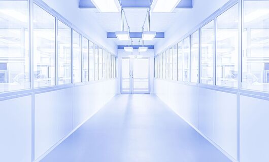 Bright white corridor with a glass door at the end. Windows on both sides reveal visible people behind them. The environment appears sterile.
