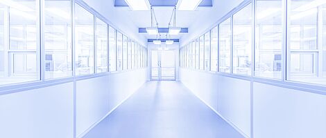 Bright white corridor with a glass door at the end. Windows on both sides reveal visible people behind them. The environment appears sterile.