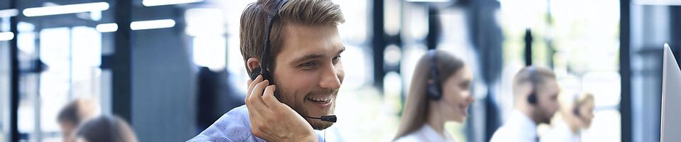 Smiling man wearing a headset sits at a desk in a call center, one hand on his ear, the other on the keyboard.
