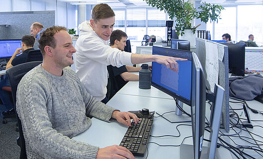 A man is sitting at a computer in an open-plan office; next to him, a young man is standing and pointing at a screen.