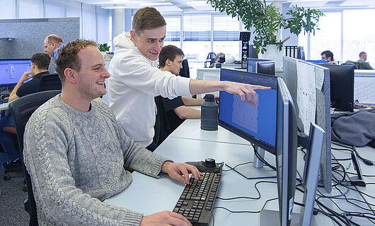 Ein Mann sitzt in einem Großraumbüro an einem PC; neben ihm steht ein junger Mann, der auf einen Bildschirm zeigt.