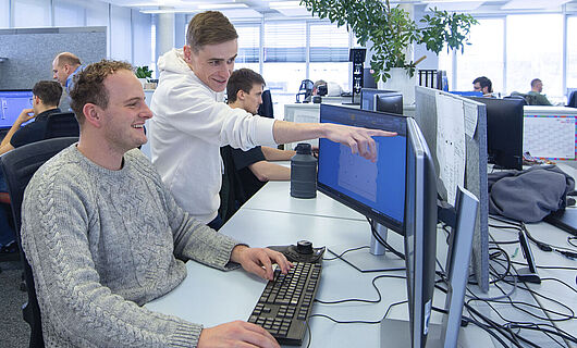 Ein Mann sitzt in einem Großraumbüro an einem PC; neben ihm steht ein junger Mann, der auf einen Bildschirm zeigt.