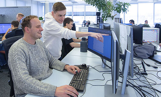 A man is sitting at a computer in an open-plan office; next to him, a young man is standing and pointing at a screen.