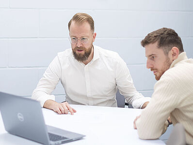 Two men are sitting in a meeting room at a table, looking together at a laptop placed on the table.