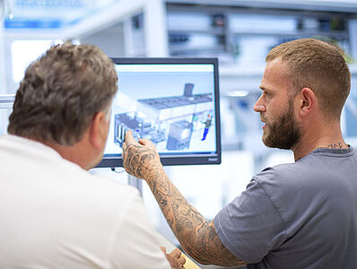 Two men with their backs to the camera are looking at a monitor displaying a machine drawing; one of them is pointing at the screen.