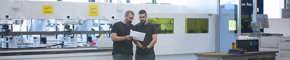 Zwei Männer stehen vor einer Tube Laser Cutting System Maschine, einer hält einen Plan in der Hand.