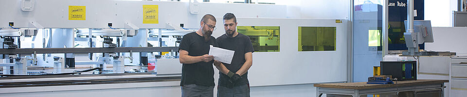 Two men stand in front of a tube laser cutting system machine, one holding a blueprint.