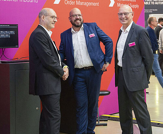 Three smiling men stand in front of a trade fair wall, engaged in conversation.