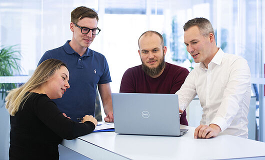 Drei Männer und eine Frau stehen gemeinsam an einem Stehtisch und schauen auf einen Laptop, der auf dem Tisch steht.
