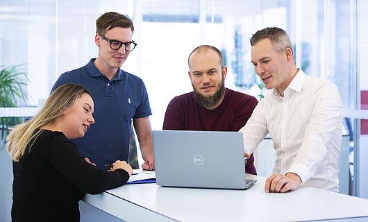 Three men and one woman are standing together at a standing table, looking at a laptop placed on the table.