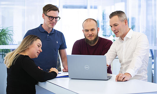 Drei Männer und eine Frau stehen gemeinsam an einem Stehtisch und schauen auf einen Laptop, der auf dem Tisch steht.