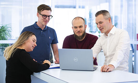 Three men and one woman are standing together at a standing table, looking at a laptop placed on the table.