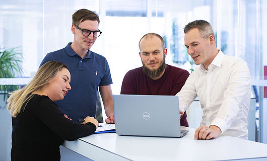 Three men and one woman are standing together at a standing table, looking at a laptop placed on the table.