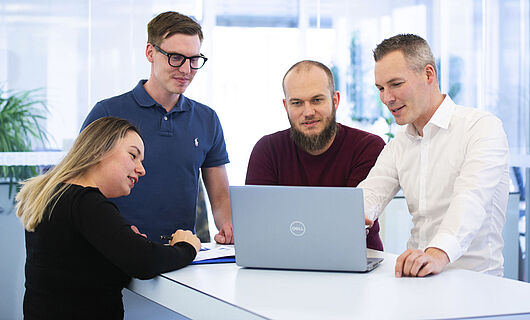 Drei Männer und eine Frau stehen gemeinsam an einem Stehtisch und schauen auf einen Laptop, der auf dem Tisch steht.