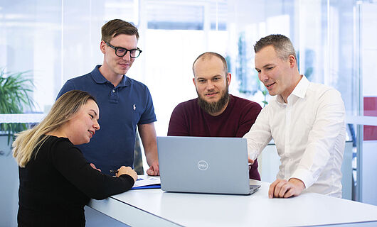 Three men and one woman are standing together at a standing table, looking at a laptop placed on the table.