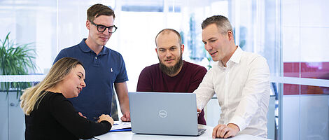 Three men and one woman are standing together at a standing table, looking at a laptop placed on the table.
