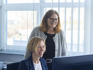 A woman is sitting at a desk in front of a computer; next to her, another woman is standing and looking at the monitor.