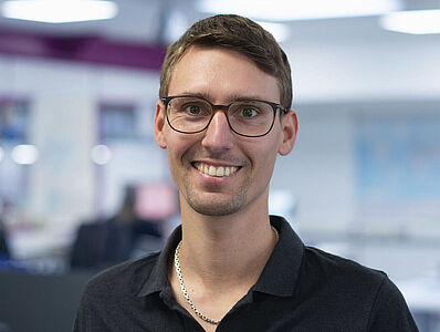  A smiling man is standing in an open-plan office.