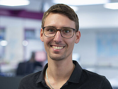  A smiling man is standing in an open-plan office.