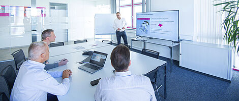 Three men are looking at a large monitor in a meeting room. One man is standing next to the monitor and giving a presentation.