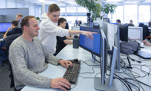 A man is sitting at a computer in an open-plan office; next to him, a young man is standing and pointing at a screen.
