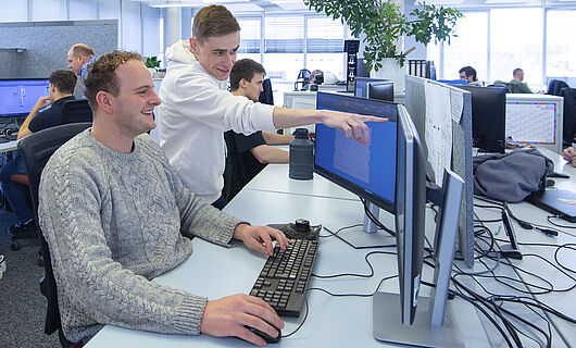Ein Mann sitzt in einem Großraumbüro an einem PC; neben ihm steht ein junger Mann, der auf einen Bildschirm zeigt.