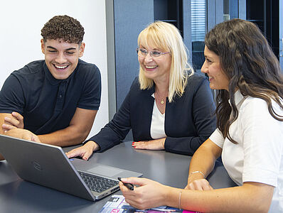 Two women and a man are sitting together at a silver table in an office, looking at a laptop.
